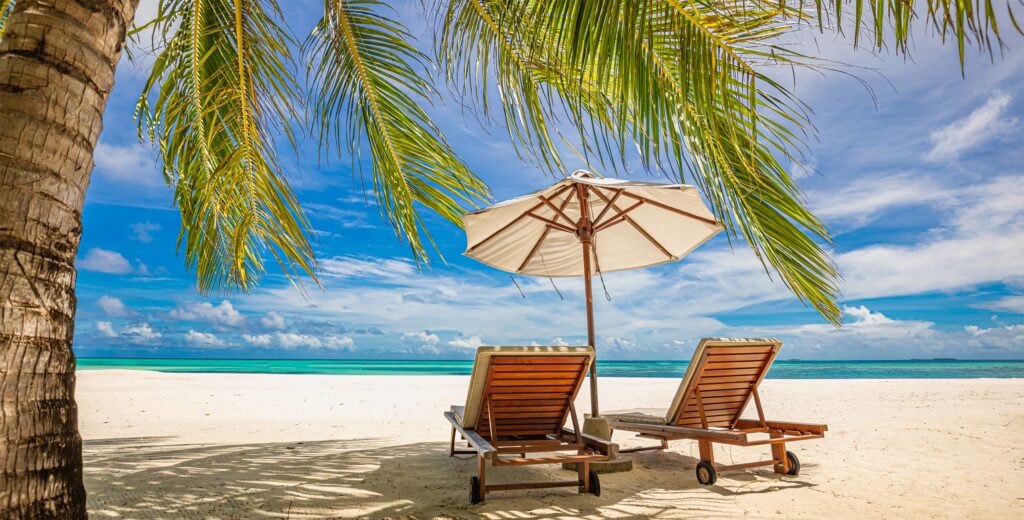 Two lounge chairs on beach with umbrella and palm tree and ocean in background